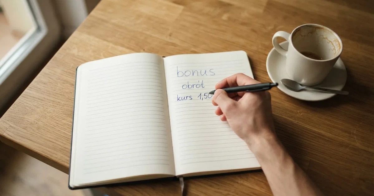 Dłoń z długopisem nad pustym notesem na kuchennym stole, poranne światło wpada przez okno.