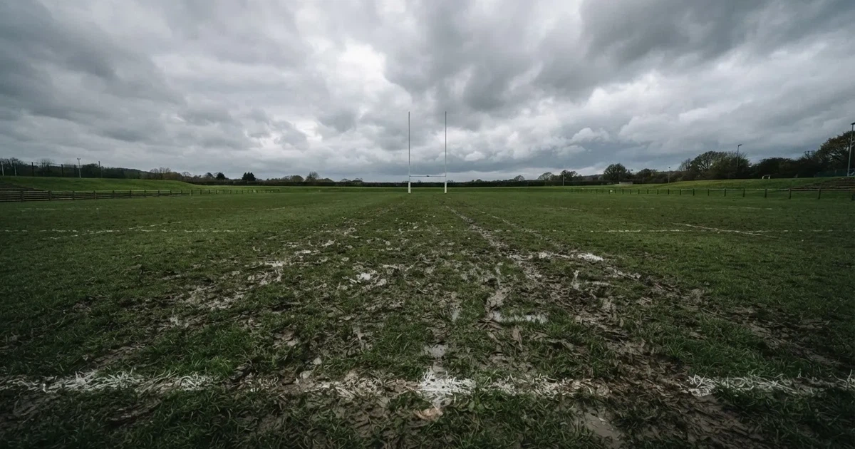 Boisko do rugby ze słupami bramki w oddali pod pochmurnym niebem, mokra trawa i ślady butów.