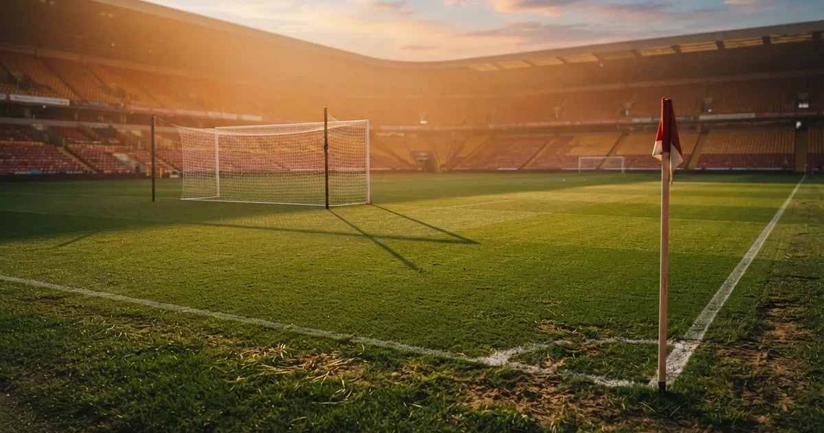Murawa stadionu o zachodzie słońca z długimi cieniami słupków bramki, trybuny w ciepłym świetle.