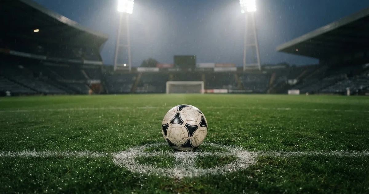 Futbolowa piłka stoi na punkcie karnym w pustym stadionie podczas mżawki, w tle rozmyte trybuny i jasne światło masztów.