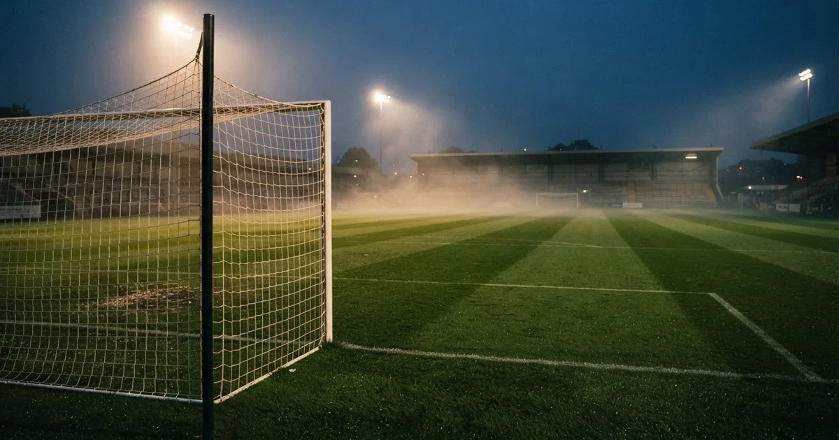 Pusta murawa stadionu miejskiego w chłodny jesienny wieczór, siatka bramki świeci w świetle reflektorów, nad boiskiem wisi mgła.