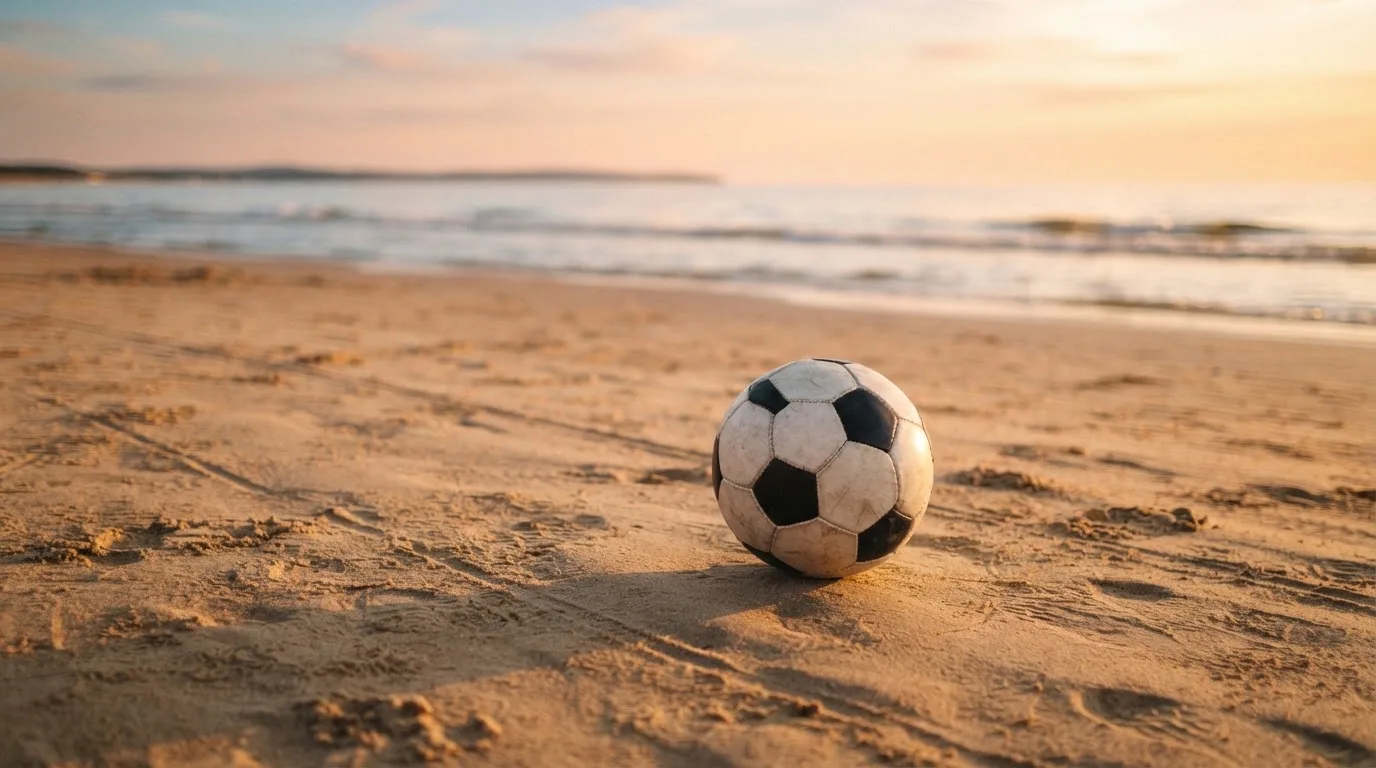 Piłka futbolowa leży na piasku boiska plażowego w ciepłym świetle zachodu, w tle morze i niski horyzont.