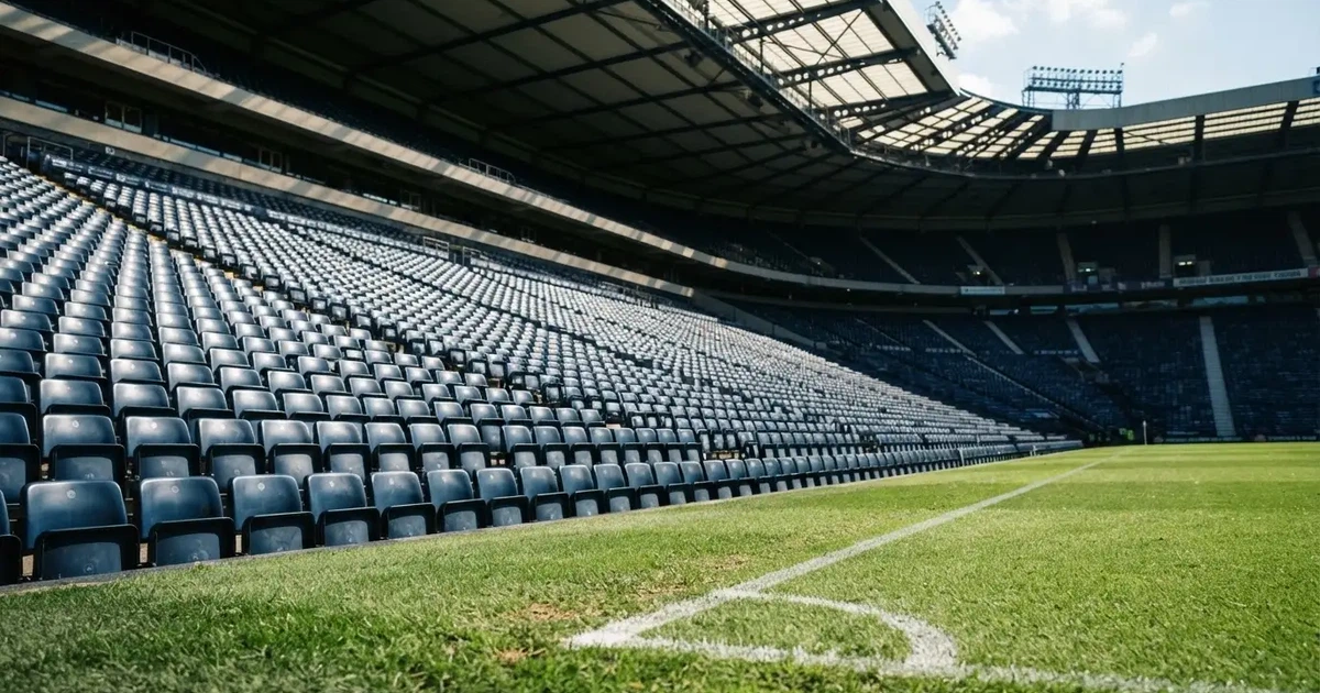 Trybuny wielkiego stadionu widziane z murawy, rzędy pustych ciemnoniebieskich krzesełek, ostre światło dnia i długie cienie.