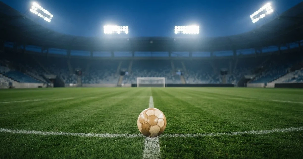 Piłka nożna leży na środku murawy dużego stadionu o zmierzchu, a nad boiskiem świecą rzędy reflektorów nad pustymi trybunami.